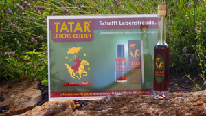 Eine Flasche mit TATAR Lebens-Elixier und ein Werbeschild vor einem Kräuterbeet. 