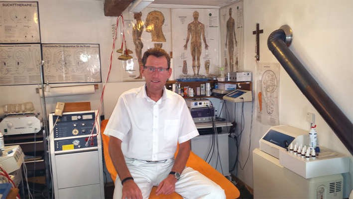 A medical practitioner (Mr. Ernst Eineder) sitting on a gurney in the treatment room of his practice.